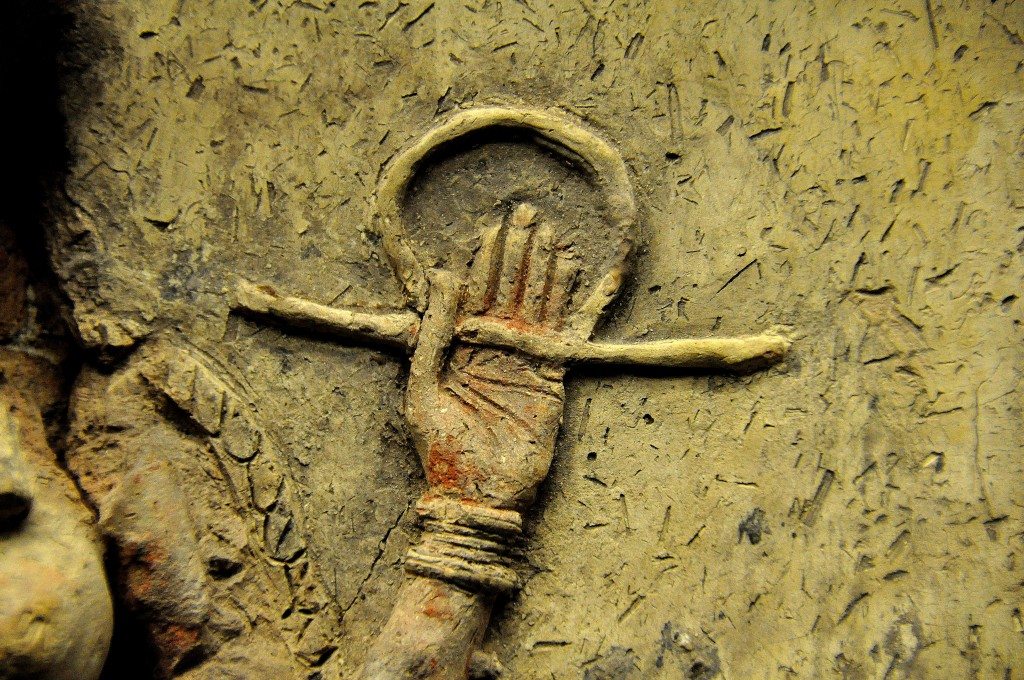 A close-up image of the left arm. The palm faces the viewer. The fingers are extended and adducted while the thumb is flexed and adducted. The hand holds the rod-and-ring symbol of divinity. Traces of red color are still seen. Note the wrist bracelet and the palm creases. 