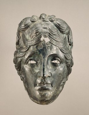 ​Mask from a Cavalry Helmet from Asia Minor, 75–125 CE. The J. Paul Getty Museum (72.AB.105). This mask was worn by a male soldier, but a Roman man would not have worn such long, carefully curled hair with a center part. The top-knot indicates it might represent Apollo, or possibly a female goddess like Aphrodite or Artemis. Because of the delicately outlined eyes and hairband, which originally included other materials such as gold, silver, or jewels, this was probably not worn in combat, but in a tournament or parade. We don’t know why the mask is shown with this hairstyle; it may relate to the military unit’s patron god or goddess, whose protection is invoked. Masks like this may also have been worn by soldiers in contests that re-enacted scenes of Greek myth and history.