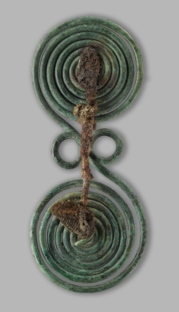 Spectacle-Shaped Brooch with Fabric Remains Early Iron Age (1000–700 BC). Copper alloy, iron, and textile. L. 5.9 in; Diam. coils 2.4 in, 2.6 in (L. 15 cm; Diam. coils 6 cm, 6.5 cm). From Olympus. Tumulus cemetery of Mesonisi, Tumulus 2, grave D. Archaeological Museum of Dion. Photo © Hellenic Ministry of Culture and Sports, Ephorate of Antiquities of Pieria, and the Dion Excavations. Courtesy Onassis Cultural Center NY.