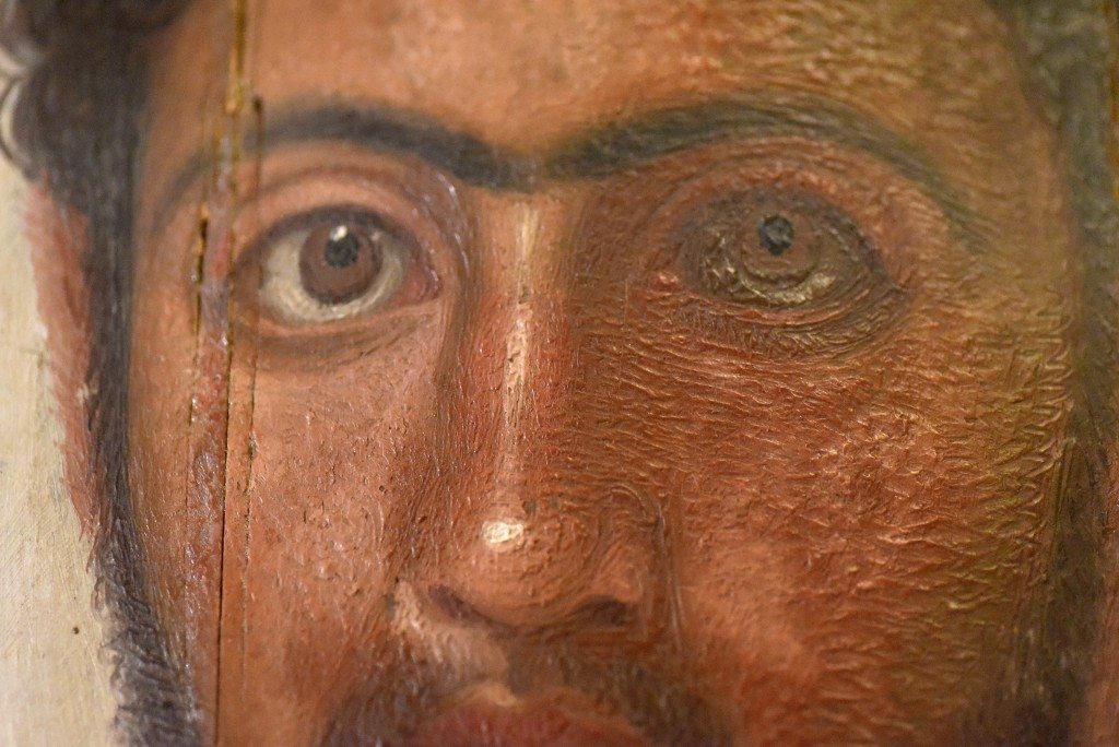 Detail of a mummy portrait from Hawara. This is a thin lime (Tilia species) wooden panel with encaustic wax mummy portrait. of Petrie's "red youth". The man has curly light beard and moustache. It appears that a sharp tool was used to delineate the shape of the face, ears, and eyebrows. From Hawara cemetery, Egypt. 1st to 2nd centuries CE. The Petrie Museum of Egyptian Archaeology, London (with thanks to The Petrie Museum of Egyptian Archaeology, UCL). The Petrie Museum has the largest collection of these "portraits" outside of Egypt. Originally, these portraits were placed over their mummified body; these images were hailed as the 1st life-like representations of real people on their 1st exhibition in London in 1888 CE. These portraits were excavated in 1888-1889 CE and 1901-1911 CE. 