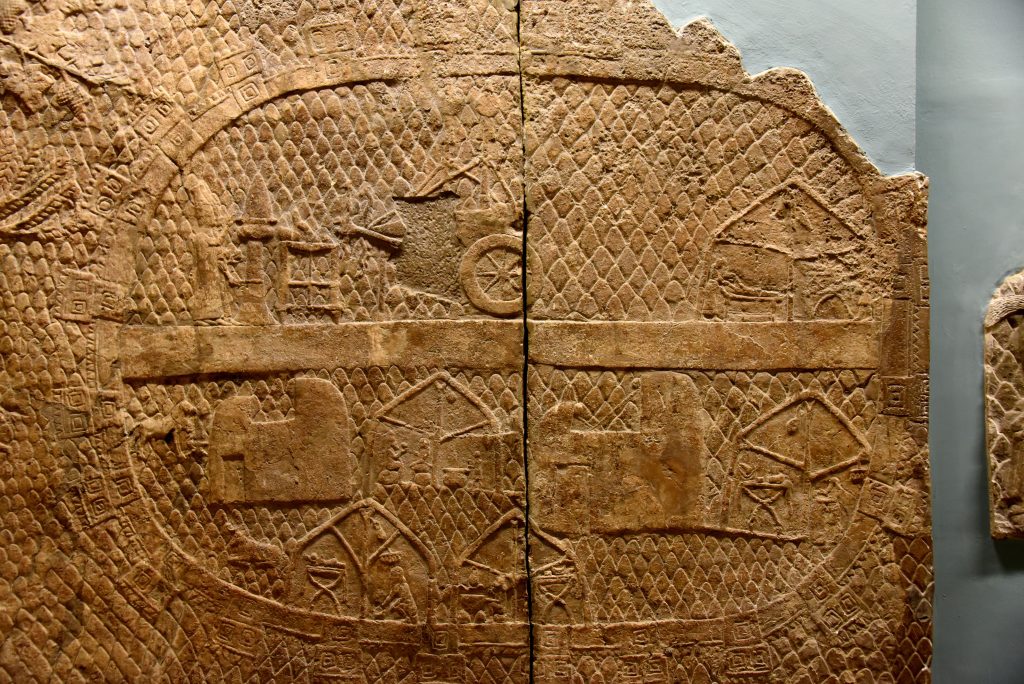 This panel shows the Assyrian base camp, from which the siege was conducted. It is fortified, with a road through the middle. Servants are work in tents, and 2 priests are performing a ceremony in front of the chariots on which are mounted the standards of the gods. Photo © Osama S. M. Amin. 