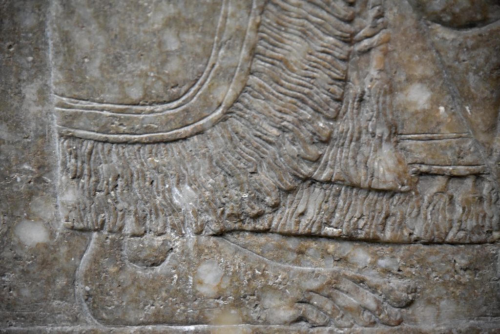 This detail of a large wall relief from Nimrud shows the lower part of the fringed wore by an eagle-headed protective spirit (Apkallu). His foot is bare. Compare this image with the similar image above, at the Sulaymaniyah Museum. The robe is different and this Apkallu stands between 2 sacred trees; there is no body before or behind him. From the North-West Palace at Nimrud, Room F, panel 8. Northern Mesopotamia, modern-day Iraq. Assyrian, 865-860 BCE. The British Museum, London. Photo © Osama S. M. Amin. 